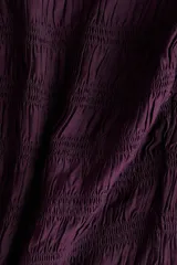 Blusa de color violeta con diseño de hombros descubiertos, cuerpo fruncido y mangas largas abullonadas con puños elásticos. Presenta detalles de volados en el escote, el bajo y los puños.