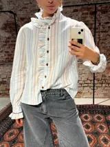 Camisa de corte clásico con estampado de rayas verticales en tonos celestes y marrones. Presenta volados delicados en el frente y en el cuello, cierre frontal con botones y mangas largas con puños ajustados.