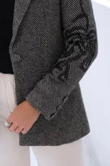 Blazer de corte sastrero en color gris con patrón de rayas diagonales y bordados negros en las mangas y solapas. Presenta cierre frontal con botón, bolsillos con solapa y estructura clásica.