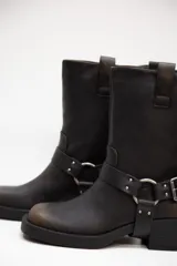 Botas de caña media en eco cuero negro con acabado mate, tacón bajo y una correa con hebilla y tachas plateadas alrededor del tobillo.