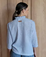 Camisa de manga tres cuartos color beige con cuello con volados y botones frontales. Presenta un detalle de color negro en la parte superior de las mangas.