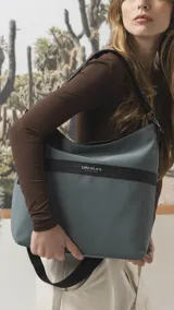 Cartera tipo hobo de tamaño grande en color beige, con detalles en negro. Presenta una franja horizontal negra con el logo de la marca, asa corta de hombro y correa larga desmontable. Incluye bolsillo frontal con cierre oculto y bolsillo trasero.