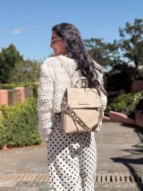 Mochila tipo cartera con tapa, confeccionada en símil cuero color suela. Cuenta con doble compartimento, división interna, cierre de seguridad y bolsillo trasero con cierre. Incluye una correa frontal decorativa con tachas metálicas.