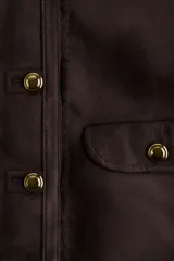 Chaqueta corta de gamuza sintética en color marrón oscuro, con forro interior de peluche. Presenta cuello redondo, cierre frontal con botones metálicos dorados y dos bolsillos delanteros con solapa y botón.