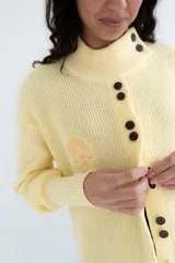 Saco tejido de punto color amarillo claro, con cuello alto asimétrico abotonado, botones oscuros contrastantes en la parte frontal y mangas largas con puños ajustados.