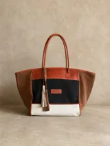 Bolso tipo shopper de gran tamaño, confeccionado en una combinación de lona y gamuza. Presenta un diseño tricolor con paneles en negro, blanco y marrón, con detalles en cuero marrón y borlas decorativas colgantes. Incluye doble asa de hombro y cierre principal.