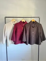 Chaqueta tipo capa de lana bouclé en color marrón, con cuello mao y cierre frontal de botones grandes. Presenta mangas tres cuartos con puños ajustados de tejido acanalado.
