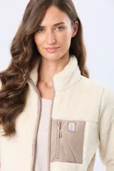 Campera de sherpa color beige con cuello alto y cierre frontal. Presenta un bolsillo frontal en el pecho de gamuza color arena con cierre y etiqueta decorativa, además de detalles en gamuza en los bordes.