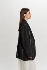 Chaqueta negra de corte oversize y diseño desestructurado, con solapas clásicas, cierre frontal de dos botones y sutil estampado de rayas verticales finas.