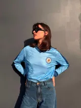 Remera de manga larga en color celeste con estampado de soles y flores de ceibo en tono azul. Presenta cuello polo y puños en tejido rib azul marino, con un parche circular bordado en el pecho que incluye el sol y la inscripción Uruguay Vdamiani.
