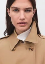 Trench coat color beige, con cuello de solapa, diseño cruzado y cierre frontal de botones. Presenta trabillas en los puños, cinturón ajustable y bolsillos laterales.