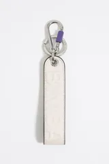 Llavero de cuero negro con la palabra "LOLA" en relieve y forro interior rosado. Incluye anilla y mosquetón metálicos dorados, con detalle de mosquetón pequeño en color fucsia brillante.