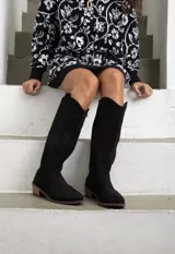 Bota de caña alta estilo western, confeccionada en gamuza negra, con taco cuadrado de madera y costuras decorativas en el empeine y la caña.