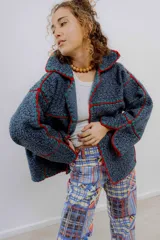 Chaqueta de corte oversize confeccionada en una fusión de paño de lana y crochet. Presenta un diseño estructurado en paneles de color azul oscuro, unidos mediante costuras decorativas visibles en hilo rojo y verde. Cuenta con cuello solapa y mangas anchas.