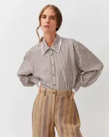 Camisa de corte recto y silueta fluida, con estampado a rayas marrones y blancas. Presenta cuello clásico, cierre frontal con botones y un bolsillo aplicado en el pecho. Las mangas largas son ligeramente abullonadas, con puños ajustados que aportan volumen a la prenda.