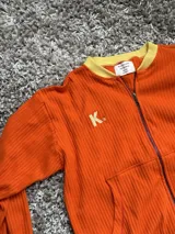 Campera de algodón premium color anaranjado, con cierre frontal, cuello alto, puños y dobladillo en amarillo. Tiene un bolsillo tipo canguro y el logo "K." bordado en el pecho.