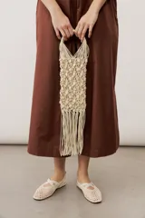 Cartera de mano de macramé color crudo con apliques de caracoles y flecos colgantes en la parte inferior. Cuenta con asas rígidas integradas en el mismo tejido.