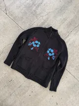 Cardigan de punto color marrón con cierre frontal, cuello alto y bolsillos aplicados. Presenta diseño tejido localizado con detalles en violeta y negro en el pecho.