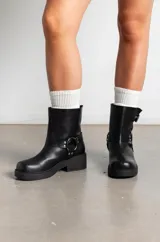 Botas de caña baja en cuero sintético color marrón, con diseño estilo biker que incluye dos correas con hebillas metálicas y una argolla decorativa en el lateral. Poseen taco bajo y suela de goma negra.