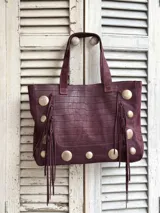 Cartera estilo shopper color bordó, confeccionada en cuero con textura tipo cocodrilo. Presenta detalles de tachas metálicas circulares y flecos laterales largos. Cuenta con doble asa de hombro.