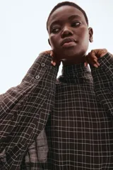 Blusa de manga larga con estampado a cuadros en tonos oscuros (negro/marrón y blanco). Presenta cuello drapeado con tira para atar en la nuca, hombros caídos y cintura entallada con pinzas moldeadoras.