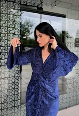 Vestido midi de fiesta en color azul, confeccionado en tela con textura satinada. Presenta un diseño cruzado con escote en V, mangas largas con puños y un detalle de fruncido en la cintura que crea una abertura frontal en la falda.