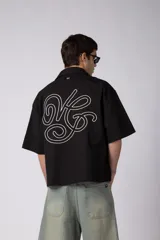 Camisa de manga corta negra 100% algodón, con calce boxy, cuello camisero, cierre frontal con botones y detalle de bordado blanco en la parte inferior.