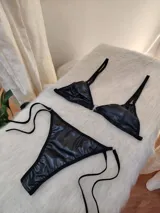 Conjunto de bikini negro con acabado efecto cuero, compuesto por un corpiño triangular con cierre frontal metálico y bombacha tipo vedetina con tiras laterales ajustables.