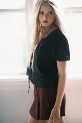 Blusa negra de viscosa con cuello Peter Pan fruncido y ribete de encaje. Presenta cierre frontal con tiras para anudar, mangas cortas abullonadas y puños con elástico.