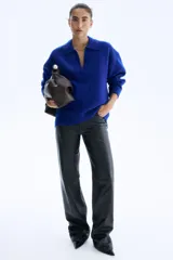 Buzo de tejido punto fino suave, color azul eléctrico, con cuello tipo polo abierto en V y hombros caídos. Presenta remates acanalados en puños y bajo.