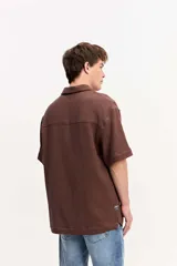 Camisa de manga corta color beige confeccionada en muselina de algodón, con cuello abierto, cierre frontal de botones y un bolsillo aplicado en el pecho.