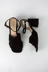 Sandalia de tacón cuadrado medio, confeccionada en material textil tipo gamuza color negro. Presenta un diseño de tiras cruzadas con nudo en la parte delantera y una tira ajustable con hebilla metálica en el tobillo.