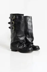 Botas de caña alta confeccionadas en cuero negro, con diseño holgado y tres correas horizontales con hebillas metálicas doradas en la parte superior. Presentan punta redondeada y taco bajo cuadrado.