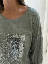 Remera de manga larga color verde, con cuello redondo y diseño holgado. Presenta un detalle frontal con parches de lentejuelas plateadas y estampado tipográfico.