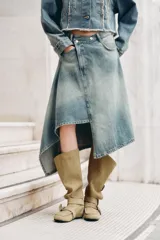 Falda midi de denim con lavado claro, tiro medio y cintura cruzada con cierre asimétrico de cremallera y botones metálicos. Presenta bolsillos delanteros y bajo asimétrico.