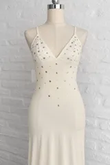 Vestido largo color marfil de corte sirena, con escote en V pronunciado, tirantes finos y aplicaciones de pedrería y tachas metálicas distribuidas en el frente.