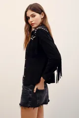 Camisa de gamuza negra con mangas largas, flecos y aplicación de tachas metálicas plateadas en la parte frontal y en los hombros.