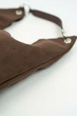 Bolso tipo hobo confeccionado en gamuza marrón, con diseño slouchy, asa corta de cuero y argollas metálicas plateadas.