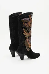 Bota de caña alta confeccionada en gamuza negra, con un diseño artesanal de parches en cueros texturizados y metalizados con motivos florales. Presenta un taco cónico y punta almendrada.