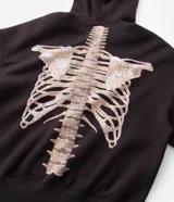 Buzo canguro marrón oscuro con capucha, cierre frontal completo y bolsillos, confeccionado en mezcla de algodón y poliéster con interior de felpa. Presenta una estampa en la espalda y mangas con diseño de huesos en tono beige claro con efecto desgastado.