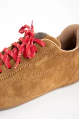 Zapatilla de estilo deportivo color camel, confeccionada en cuero gamuzado. Presenta cordones rojos con detalles blancos y un pequeño refuerzo gris en el talón.