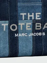 Cartera tipo tote de Marc Jacobs, confeccionada en denim con diseño de paneles en diferentes tonos de azul. Presenta el logo de la marca estampado en el frente, asas superiores de mano y una correa de hombro ajustable y extraíble en color azul oscuro.