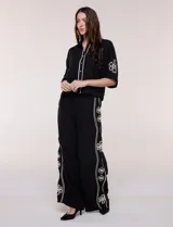 Conjunto de dos piezas negro compuesto por camisa de manga corta con cuello camisero y pantalón palazzo de tiro alto. Presenta detalles de bordados florales en contraste a lo largo de los laterales del pantalón y en las mangas.