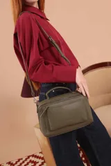 Bandolera de diseño rectangular en color suela, confeccionada en material sintético. Cuenta con un asa superior fija, correa larga ajustable y extraíble, y dos bolsillos frontales con cierre metálico, uno de ellos con diseño superpuesto. Presenta el logo de la marca grabado en el frente.