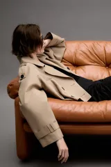 Trench corto de color beige con cuello de solapa, cierre frontal cruzado con botones y mangas largas con trabillas ajustables en los puños. Presenta un diseño estructurado con hombreras y bolsillos delanteros de vivo.