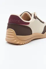 Zapatilla urbana de mujer con silueta retro, combinando paneles de cuero blanco y gamuza gris claro. Posee un talón trasero en gamuza negra y suela robusta de goma color caramelo con relieve.