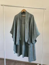 Ruana de diseño estructurado confeccionada en tela tipo tapiz, con caída fluida y corte abierto. Presenta un tono verde azulado con textura sutil.