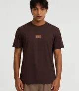 Remera de algodón peruano color marrón oscuro, corte comfort, con cuello redondo y mangas cortas. Presenta un pequeño parche rectangular estampado en el pecho con texto en tonos naranja y blanco.