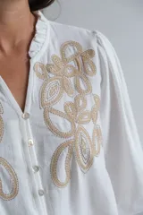 Blusa blanca de corte holgado con cuello mao y cierre frontal de botones. Presenta mangas largas abullonadas con puños elásticos y un delicado bordado en color beige en la parte superior delantera.