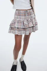 Pollera mini a cuadros en tonos tierra, azul marino y blanco. Diseño de tiro alto con elástico revestido en la cintura y cordón ajustable. Presenta dos volados horizontales que le dan volumen.